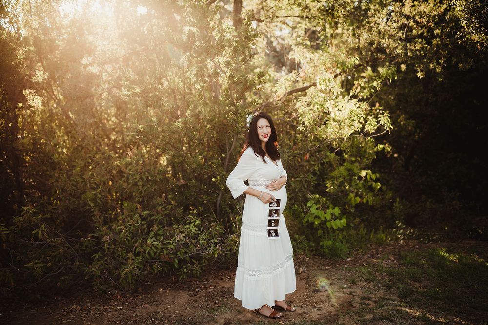 Pregnant woman in white dress standing in sunlit forest clearing, cradling baby bump with hands.