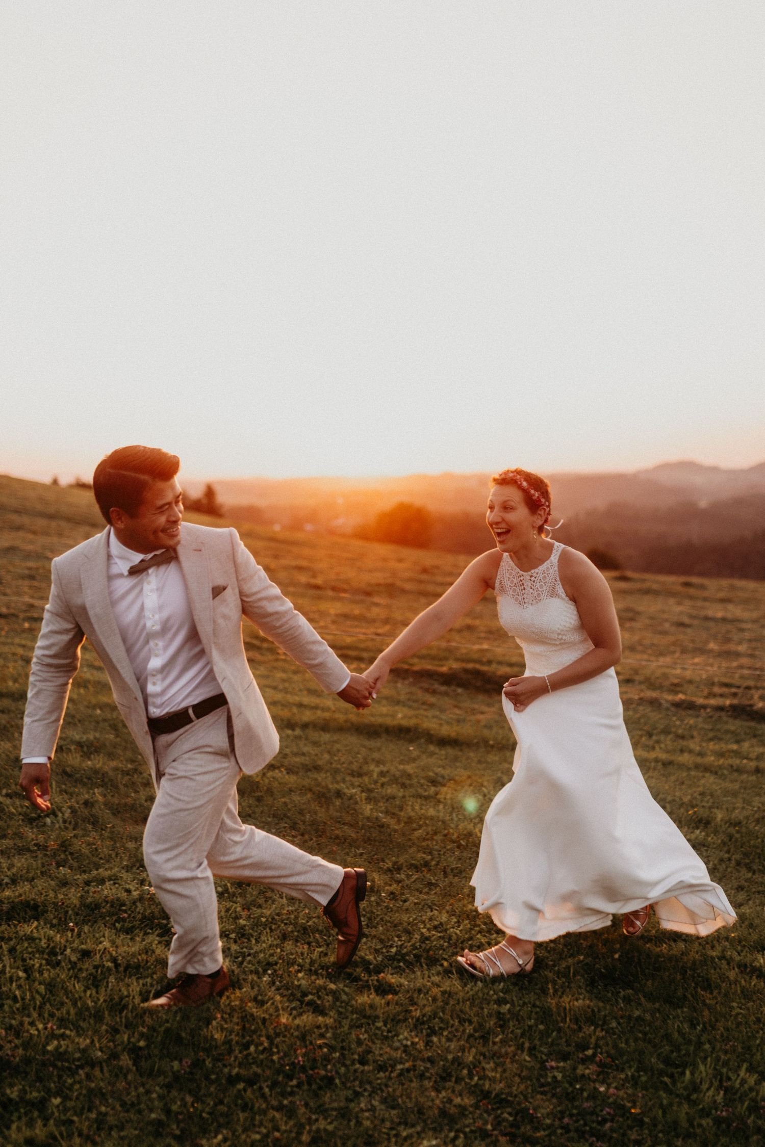 Joyful bride and groom holding hands running through green meadow at golden sunset with mountain views.