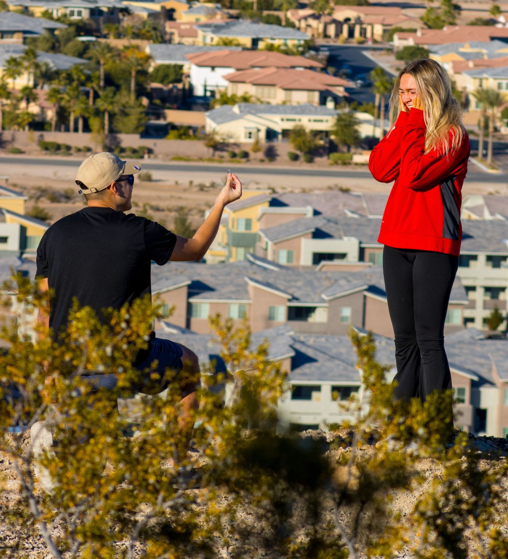 Man proposes to surprised blonde woman on hilltop overlooking Las Vegas suburban neighborhood.