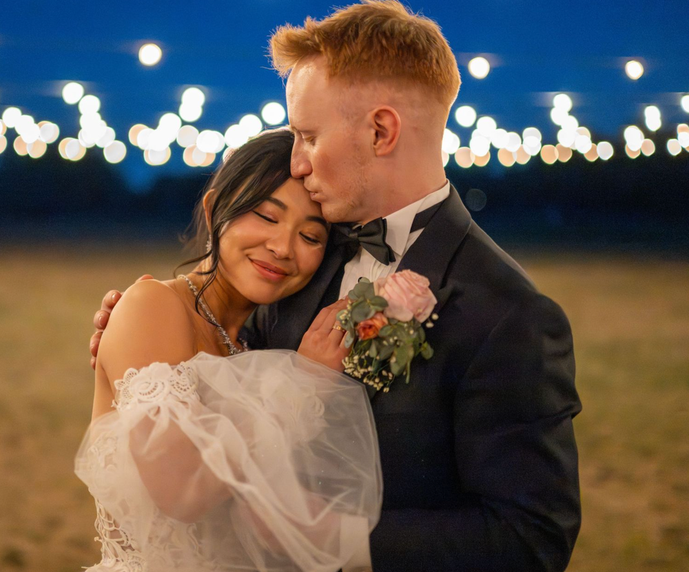 A groom in a navy suit kisses his bride in a white dress under string lights at an outdoor evening wedding.