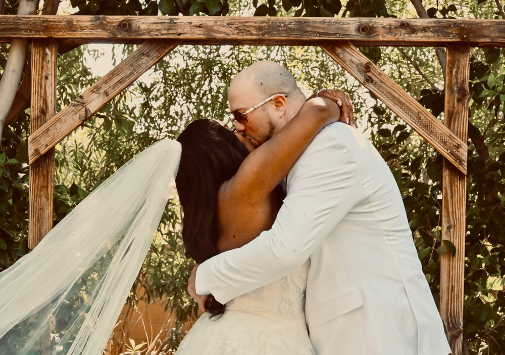 Couple sharing first kiss under rustic wooden arch at outdoor wedding ceremony.