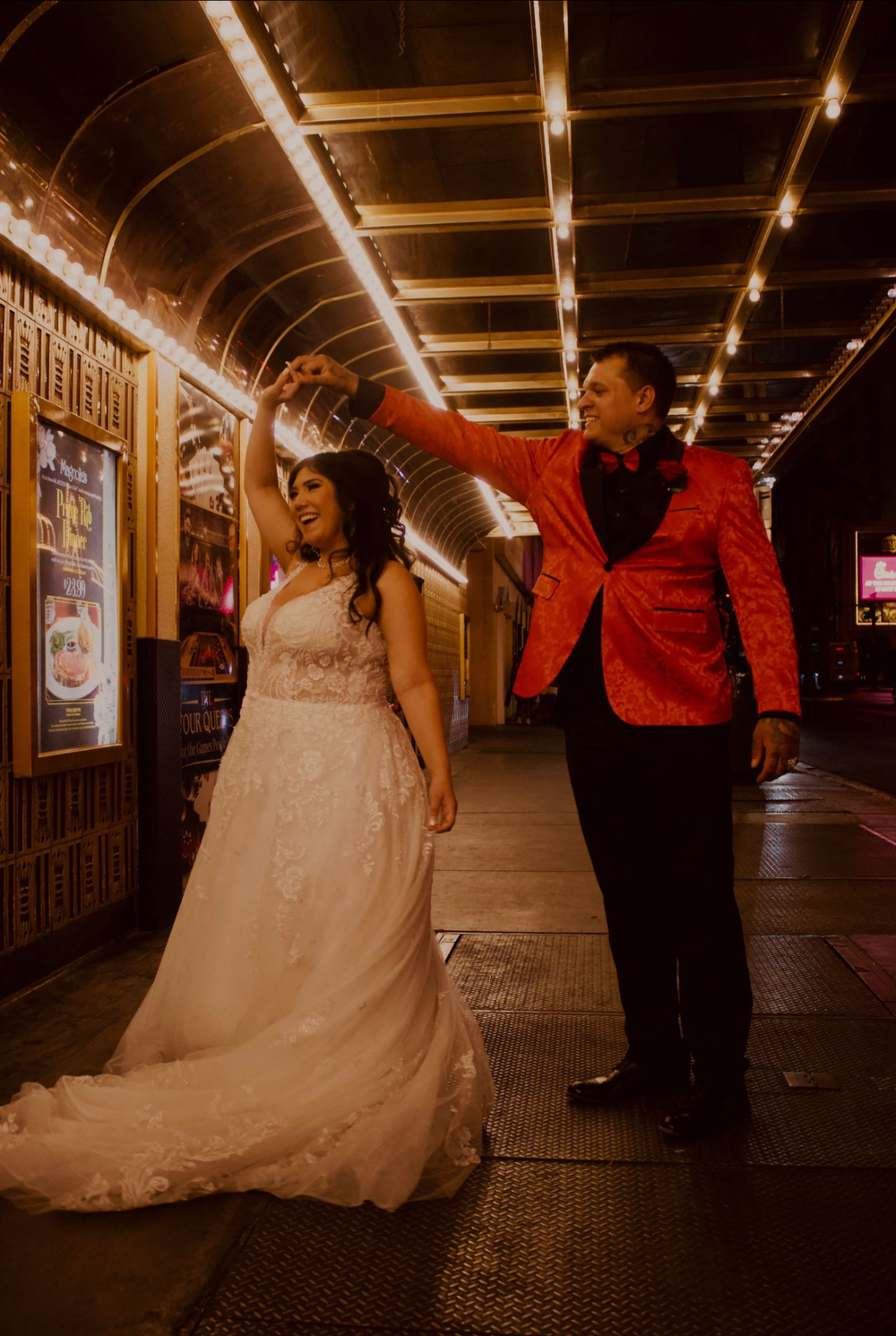 Bride in white gown twirls as groom in red sequin blazer leads her under marquee lights on a city street.