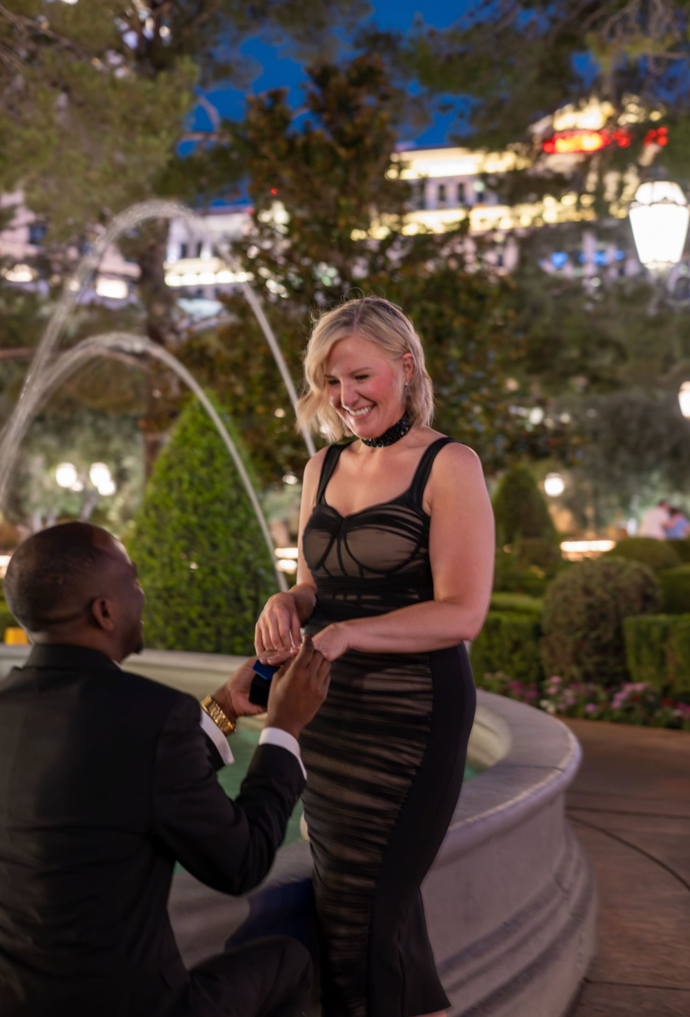 A man kneeling on one knee proposing to a smiling blonde woman in a black dress near a fountain at night.