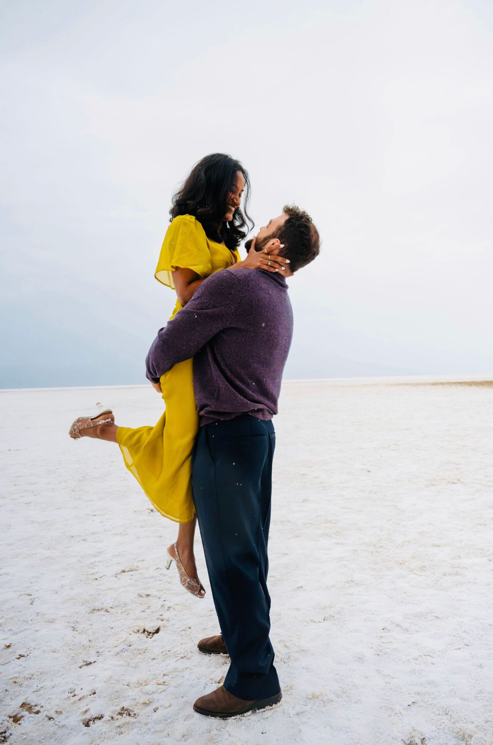 A man lifts a woman in a yellow dress on white salt flats, sharing a romantic moment under a cloudy sky.