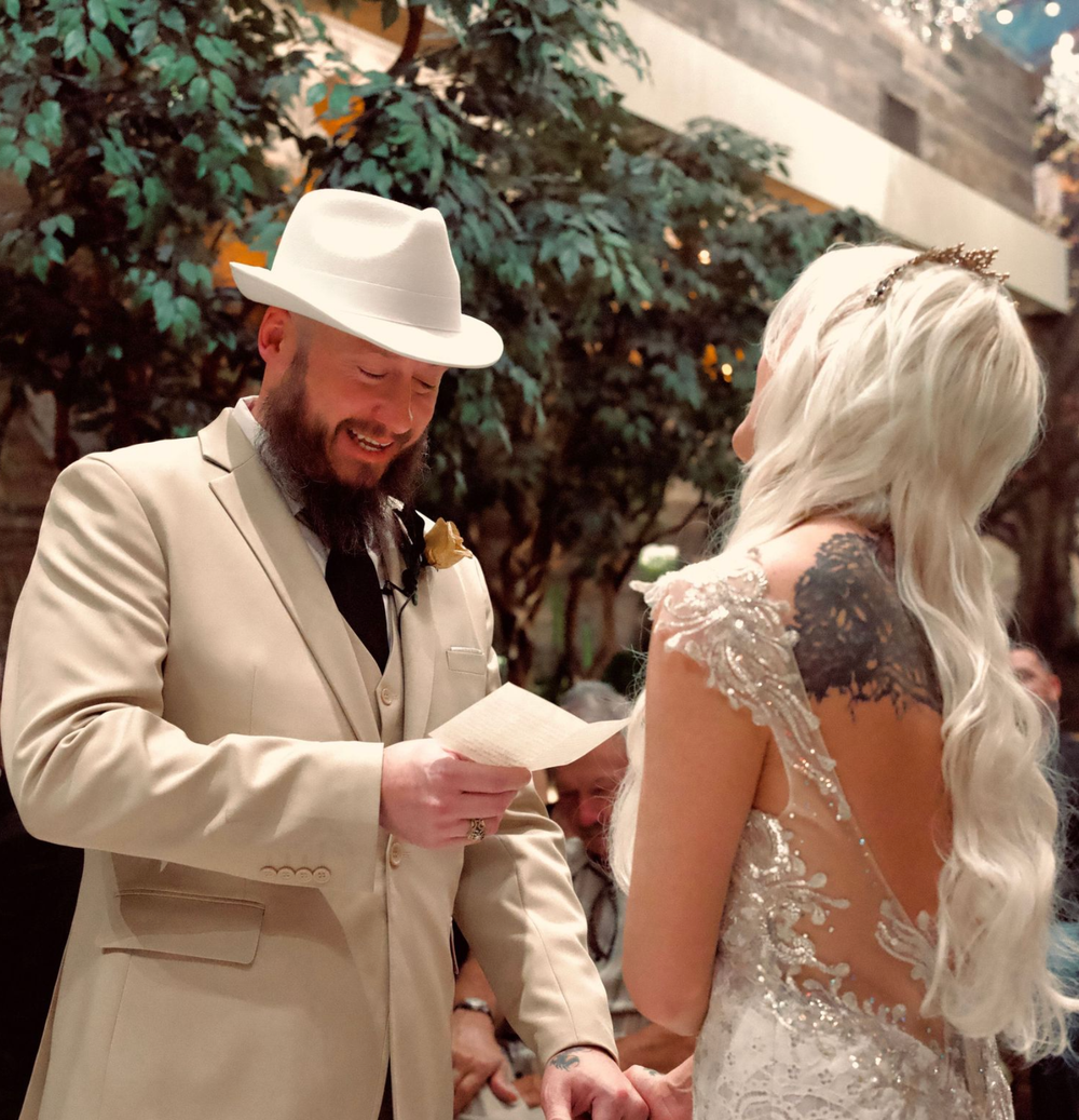 Groom in tan suit and white cowboy hat reads vows to bride in lace backless wedding dress at indoor ceremony.
