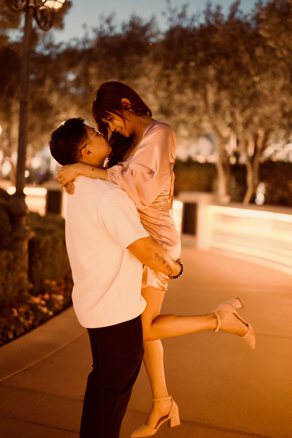 A couple shares a romantic moment at night, as a man lifts a woman in his arms near softly lit trees.
