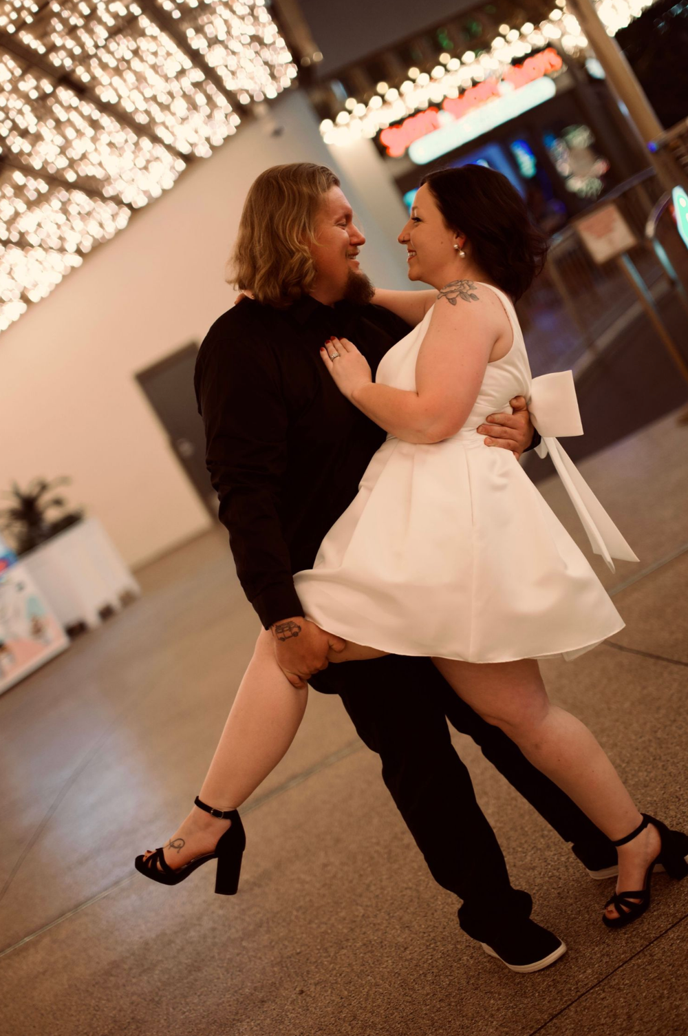 Couple dancing outdoors at night in wedding attire, woman in white dress and man in black, with bokeh lights in background.