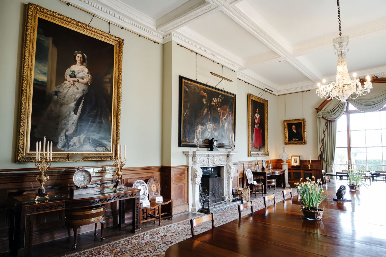 Elegant Georgian drawing room with ornate fireplace, oil paintings, Persian rug, and classic furniture in soft green tones.