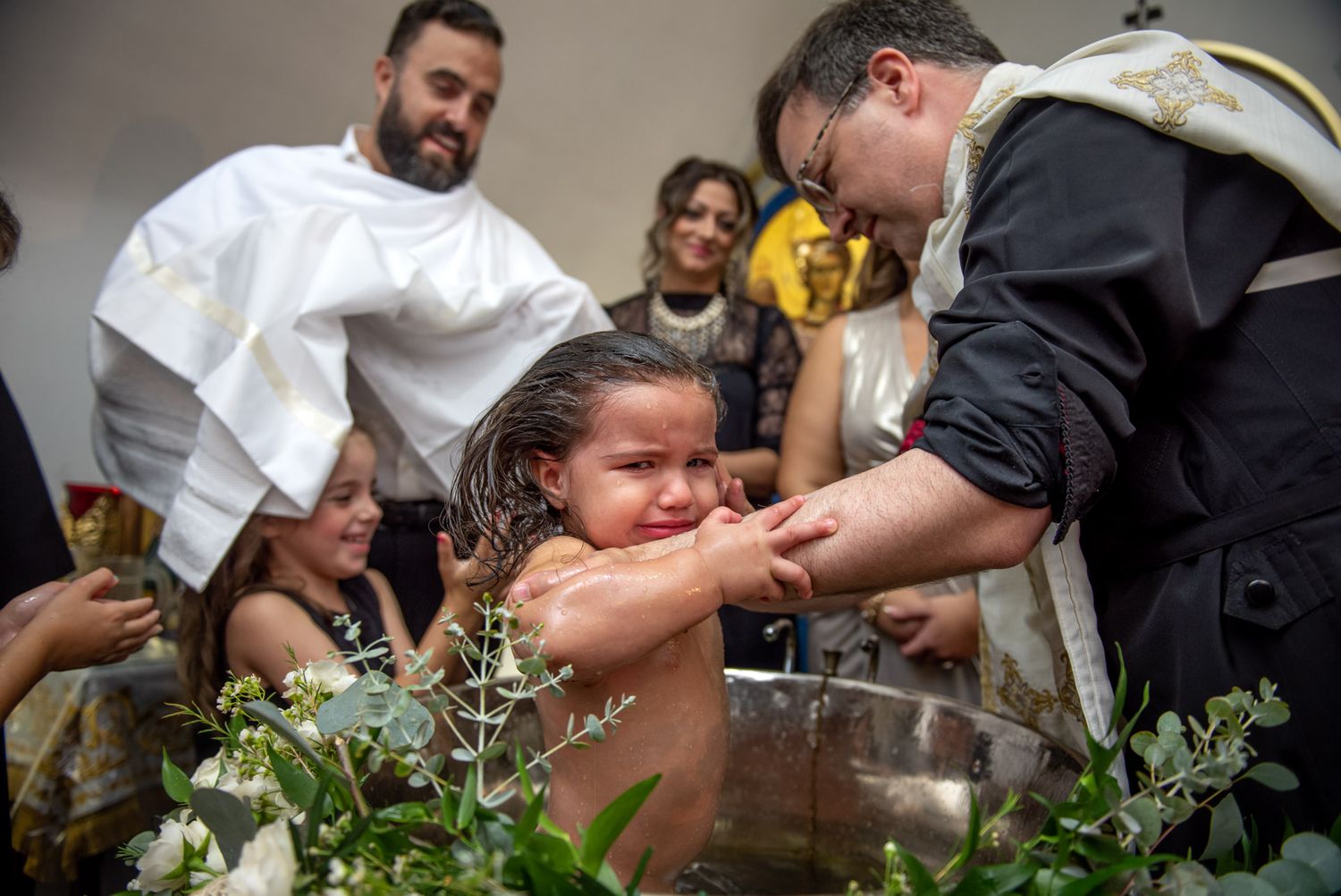 Young child being baptized in Orthodox Christian ceremony with priest and family gathered around.