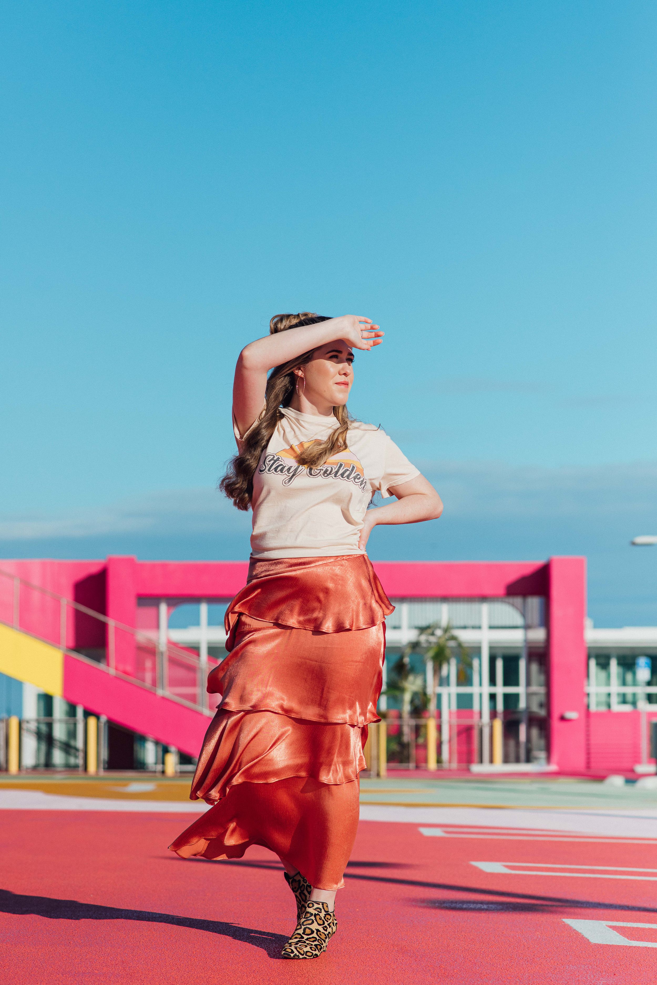 Young woman in Stay Golden tee and orange ruffled skirt poses on a vibrant pink rooftop under blue sky.