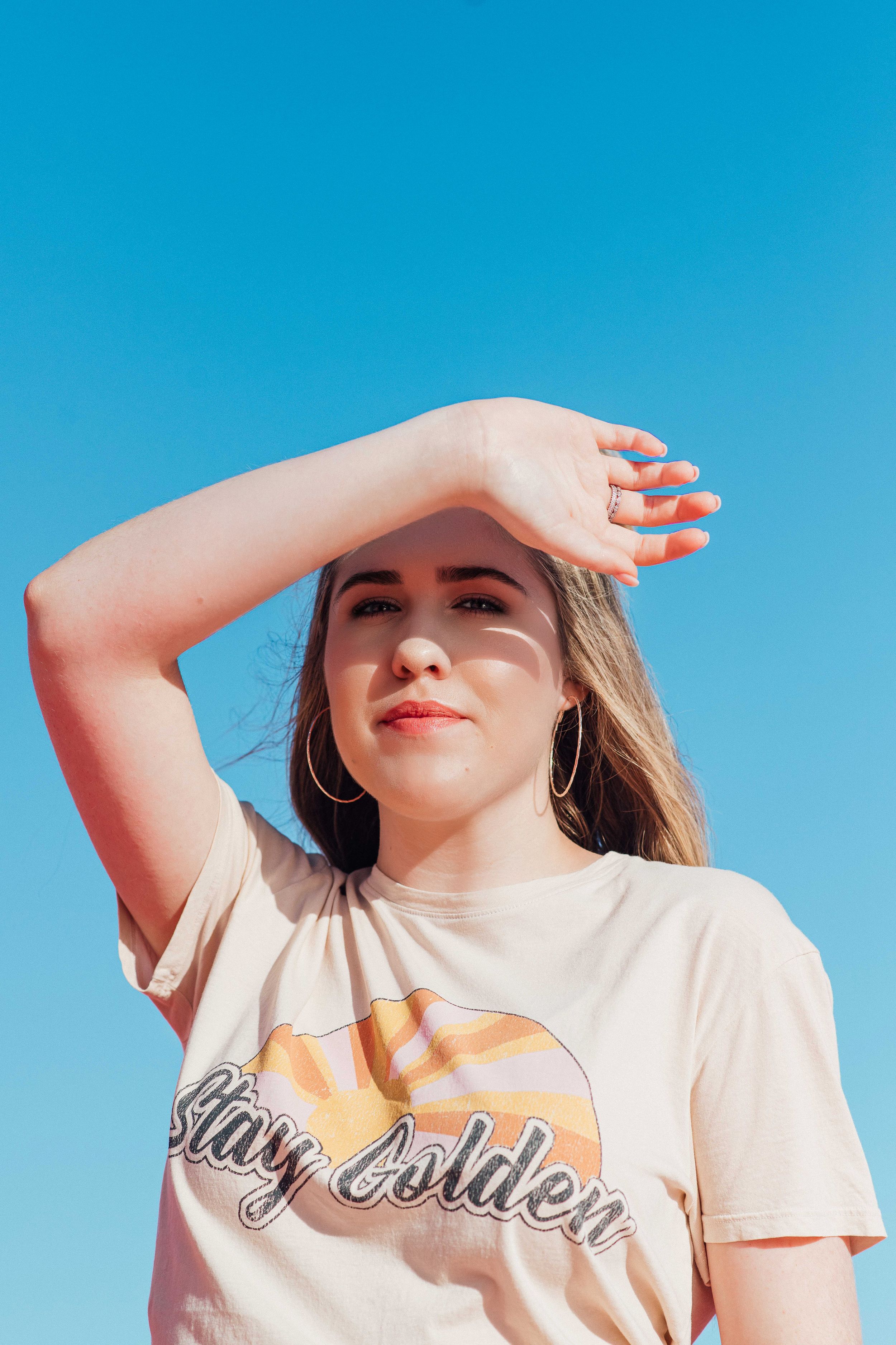 Young woman in Stay Golden graphic tee shields her eyes from the sun against a clear blue sky.