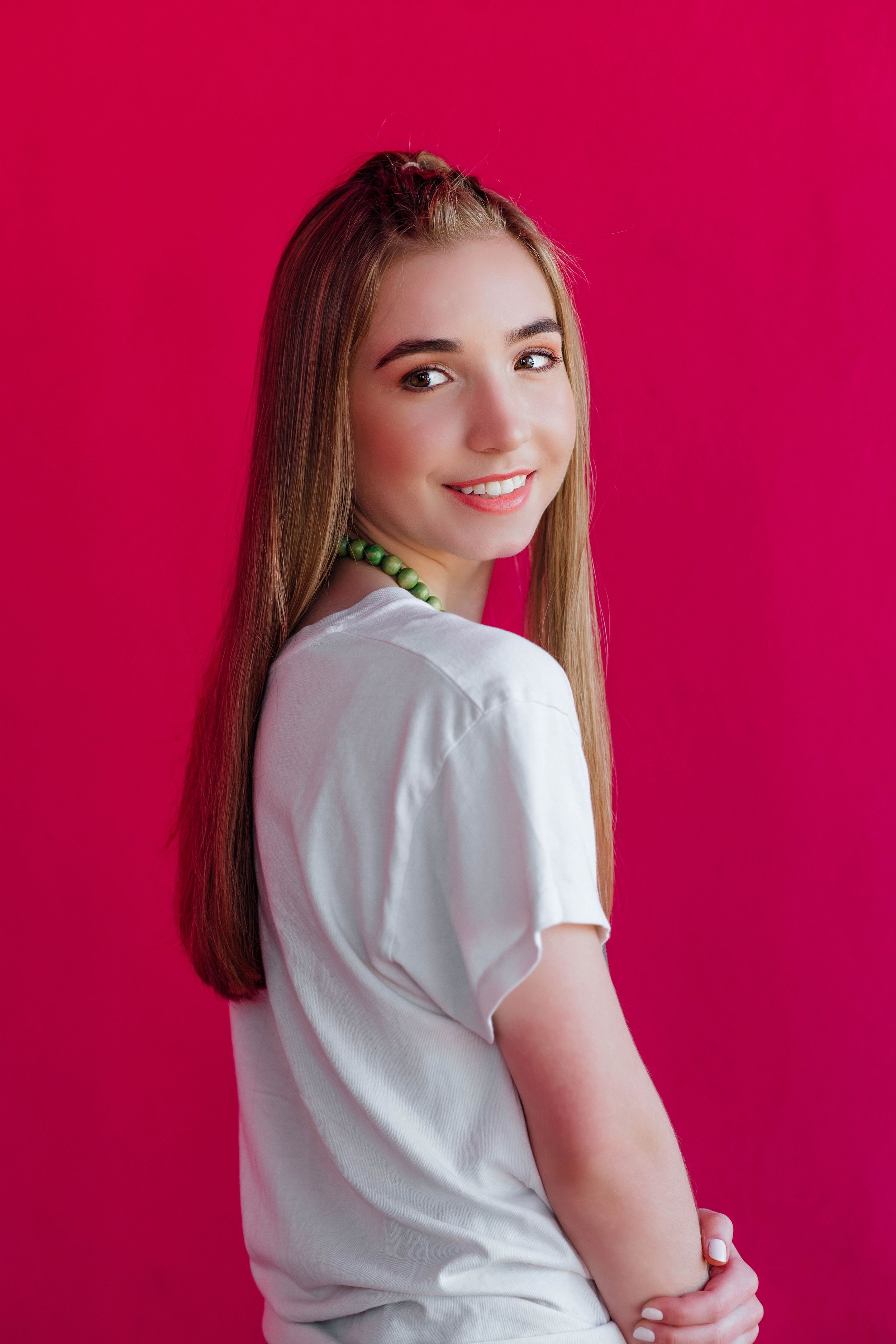 Smiling teenage girl in white t-shirt and green necklace poses against a vibrant pink background.