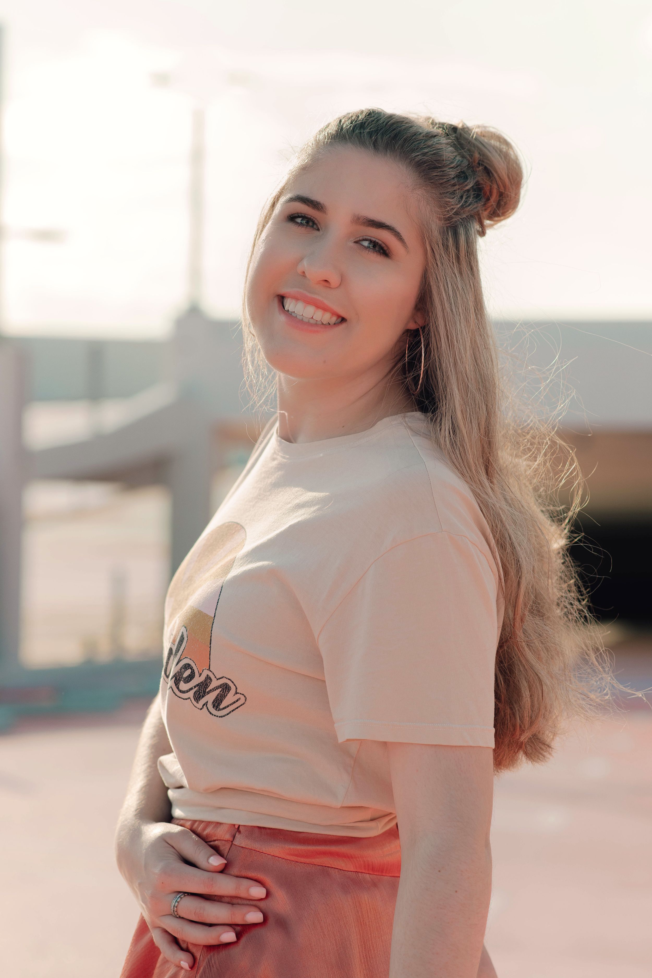 Smiling young woman in peach graphic tee with half-up bun poses in warm golden sunlight outdoors.