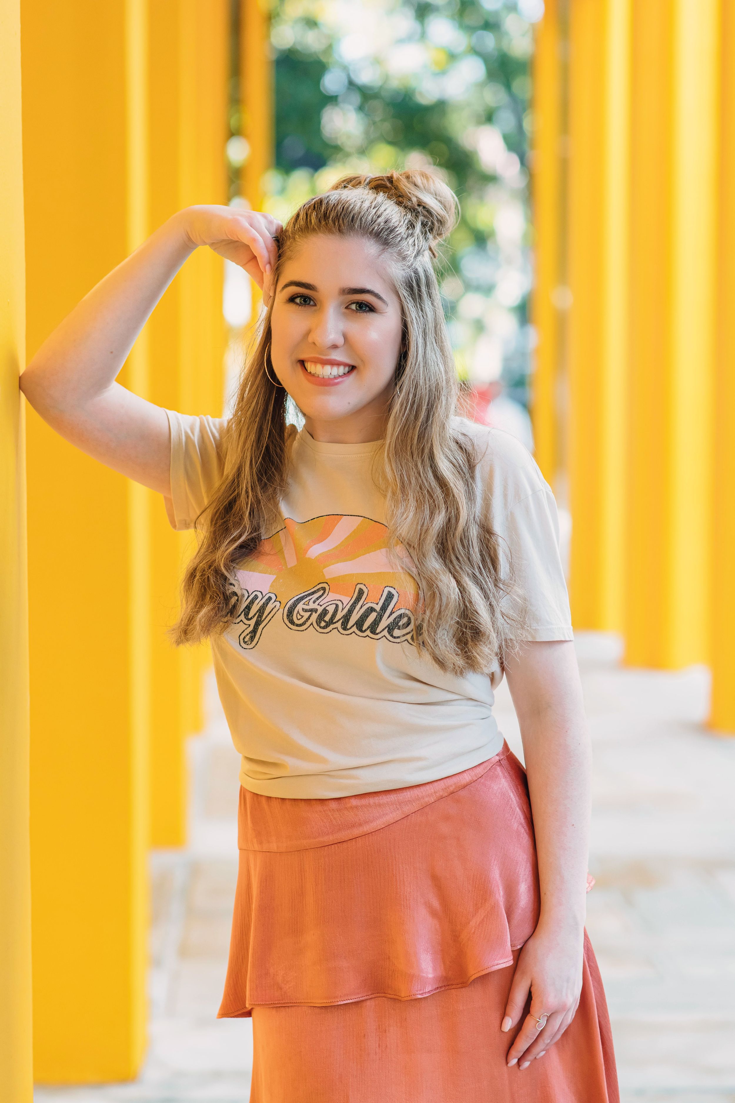 Smiling blonde woman in a 'Stay Golden' tee and pink ruffle skirt poses between yellow columns.