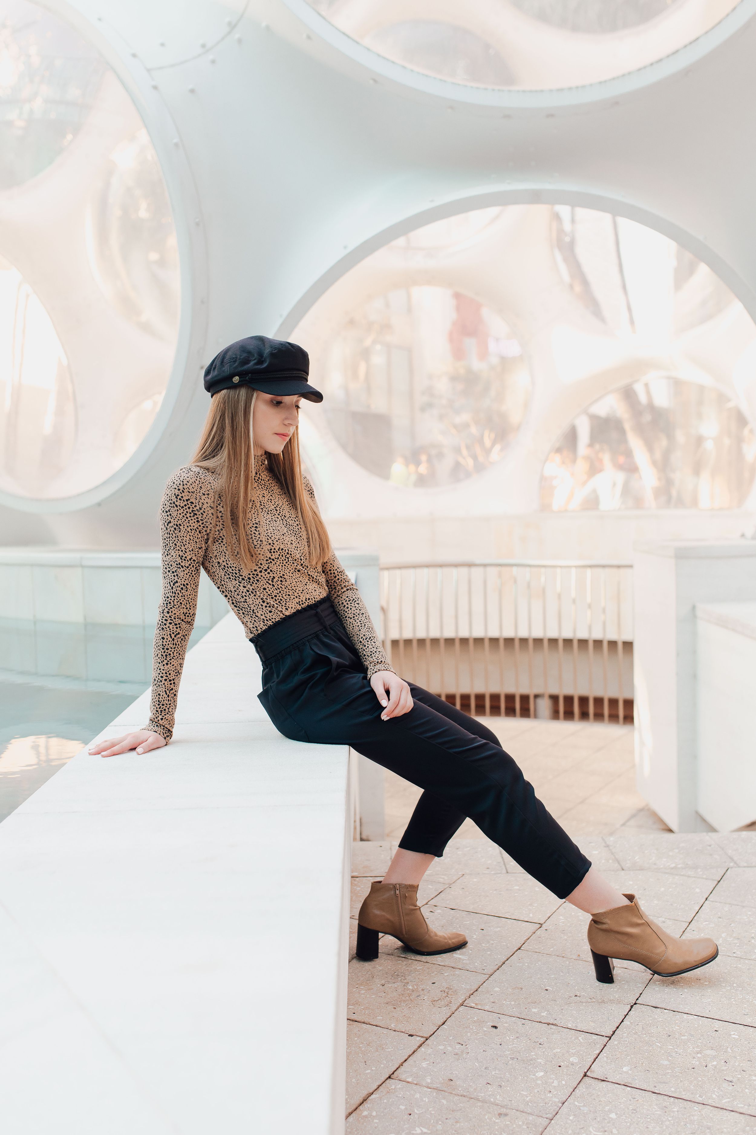 Stylish woman in leopard print top, black trousers, and cap leans on a white ledge indoors.