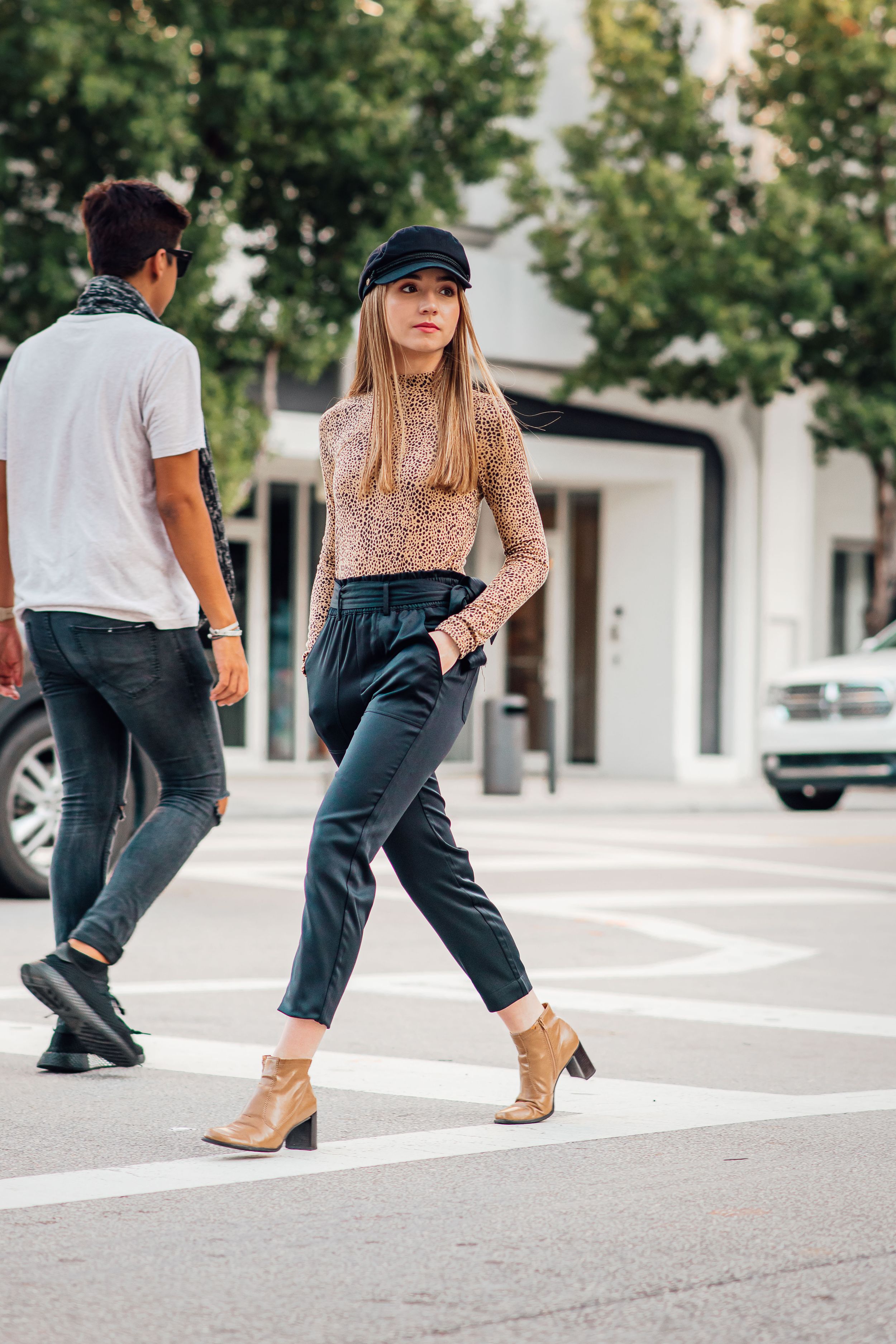 Fashionable blonde woman in leopard print top and black trousers crosses a city street crosswalk.