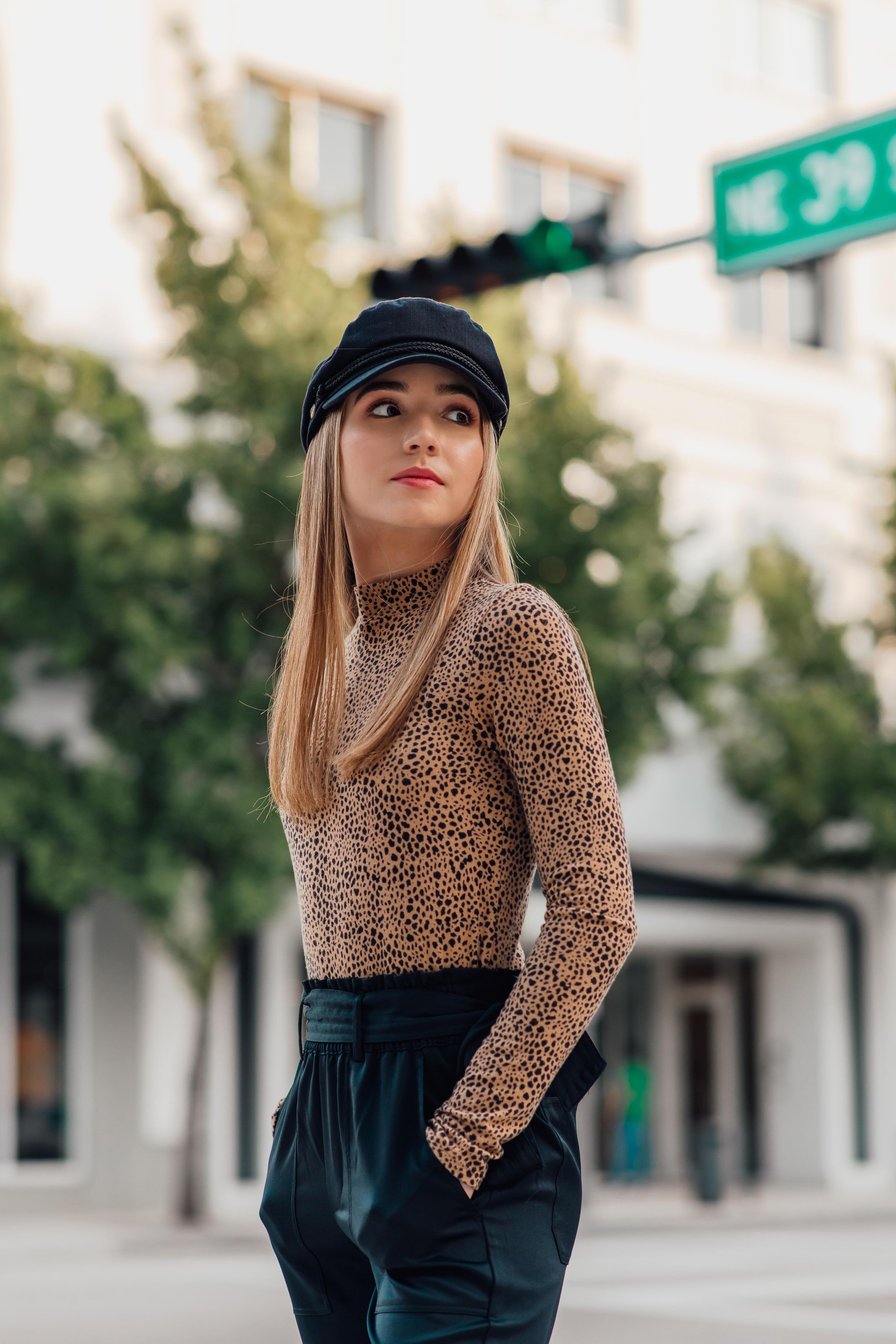 Blonde woman in leopard print turtleneck and black baker boy cap poses on an urban street corner.