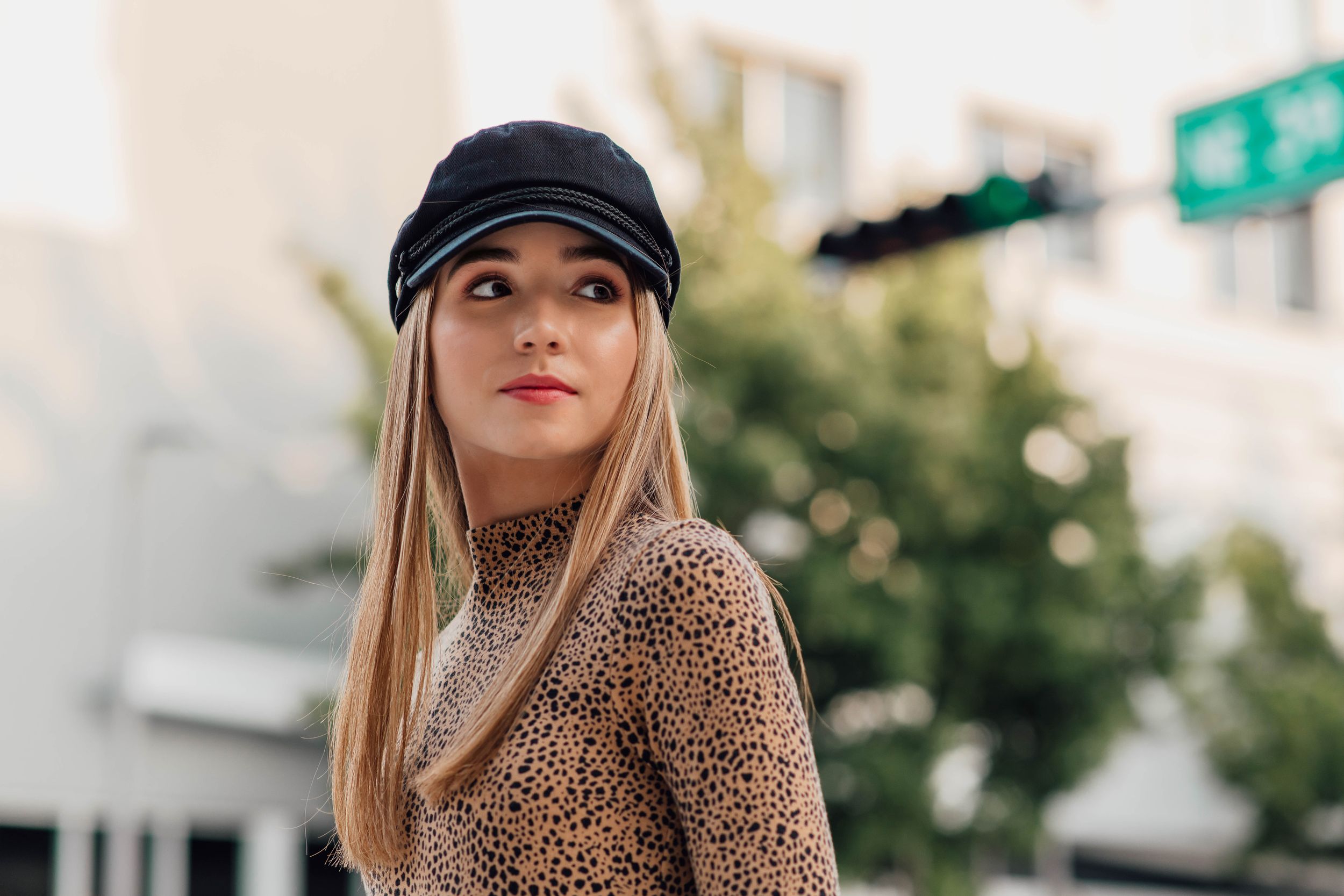 Close-up of a blonde woman in a black baker boy cap and leopard print top on a city street.