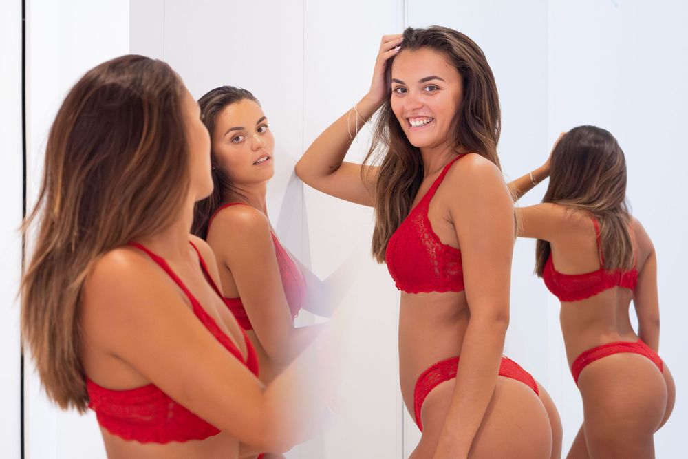 Woman in red lace lingerie set smiling at mirror reflection in bright white room.