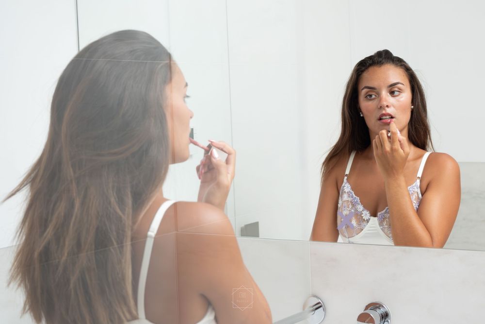 Young woman applying lipstick while looking at her reflection in a bathroom mirror.