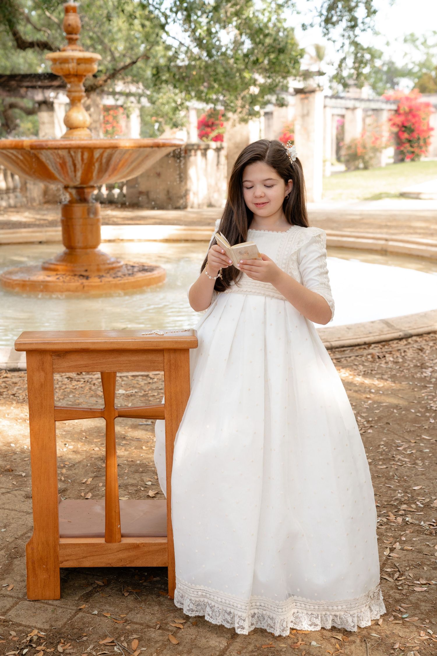 Young girl in white First Communion dress reading a book by a stone fountain in a garden.