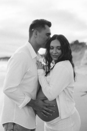 Black and white beach maternity photo of couple embracing, man kissing woman's temple while cradling her baby bump.