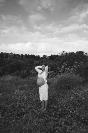 Black and white photo of a pregnant woman in a white dress standing in a field, looking up at a cloudy sky.