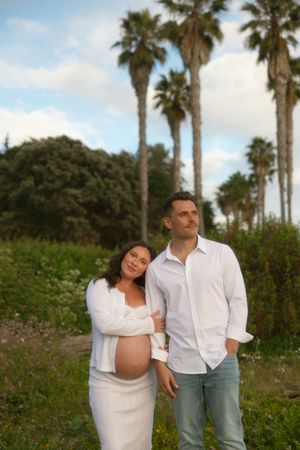 Pregnant woman in white dress leans on man in white shirt surrounded by palm trees outdoors.