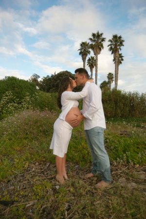 Pregnant couple kissing outdoors near palm trees, woman in white dress cradling baby bump, man in white shirt and jeans.