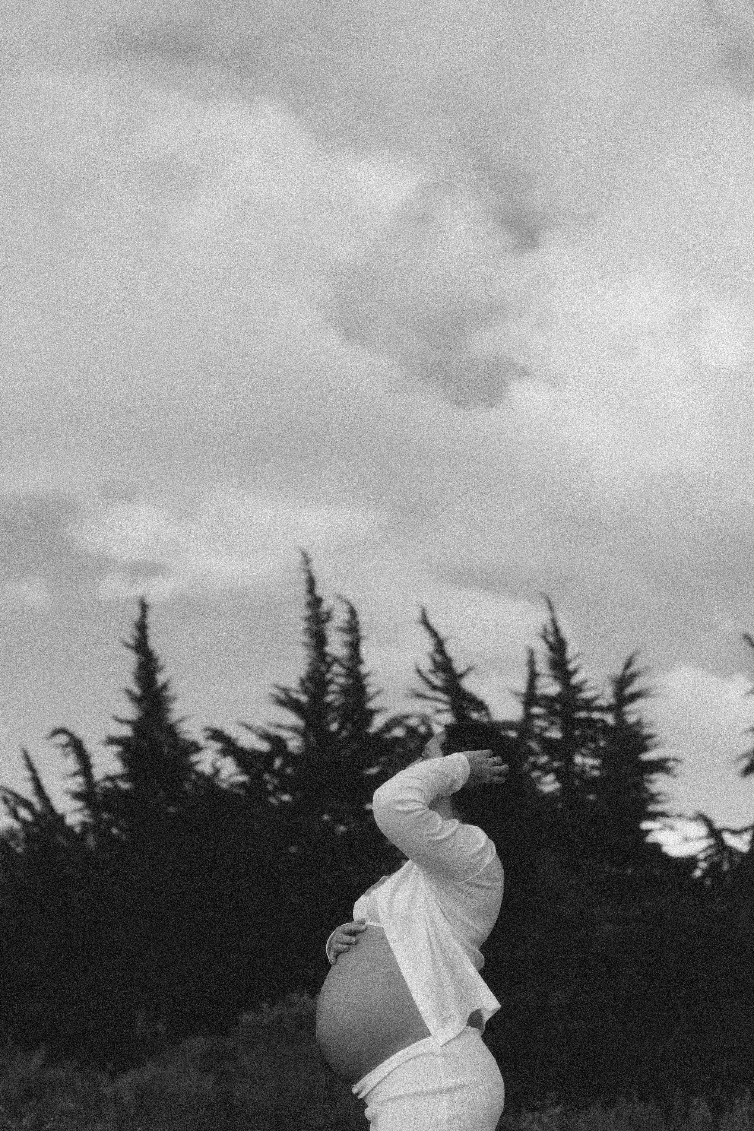 Pregnant woman in white dress poses outdoors in black and white photo with evergreen trees and cloudy sky.
