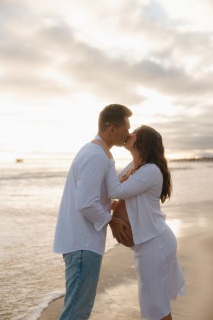 Couple sharing a romantic kiss on the beach at sunset, woman in white dress showing baby bump.