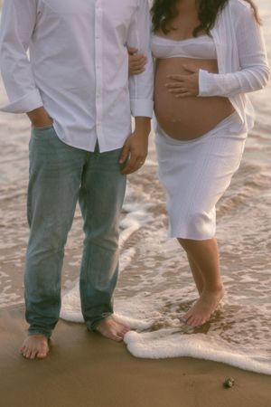Pregnant couple standing barefoot on beach shoreline, woman in white dress cradling baby bump, waves washing over feet.