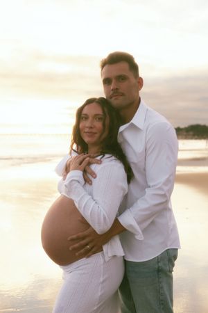Couple embraces on beach at sunset, pregnant woman in white dress held by man in white shirt.