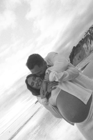 Black and white photo of a couple embracing romantically on a beach, man kissing pregnant woman's cheek by the ocean.