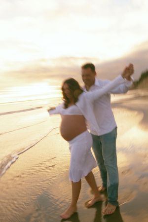 Expectant couple dancing joyfully on sandy beach at golden sunset, woman in white dress showing baby bump.