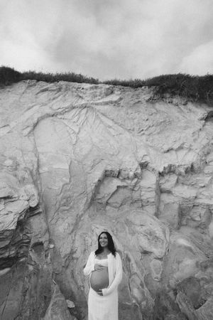 Smiling pregnant woman in white dress stands before large sandy cliff face in black and white photo.