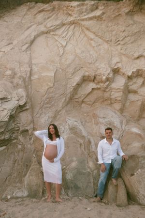 Pregnant couple in white posing against textured sandstone rock wall, maternity photo shoot on beach.