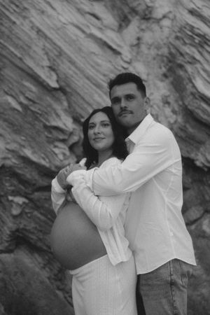 Black and white maternity photo of couple embracing against rocky background, pregnant belly visible.