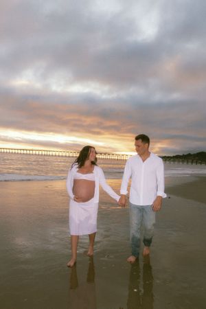 Pregnant couple holding hands walking on beach at sunset, woman in white dress showing baby bump near pier.