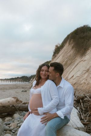 Pregnant couple sitting on driftwood at beach, man in white shirt kissing woman's cheek as she cradles her bump.