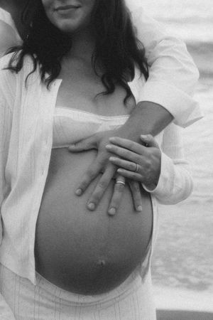 Black and white maternity photo of pregnant woman with partner's hands on baby bump, both wearing wedding rings.