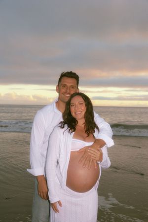 Smiling couple posing on beach at sunset, pregnant woman in white outfit cradling baby bump.