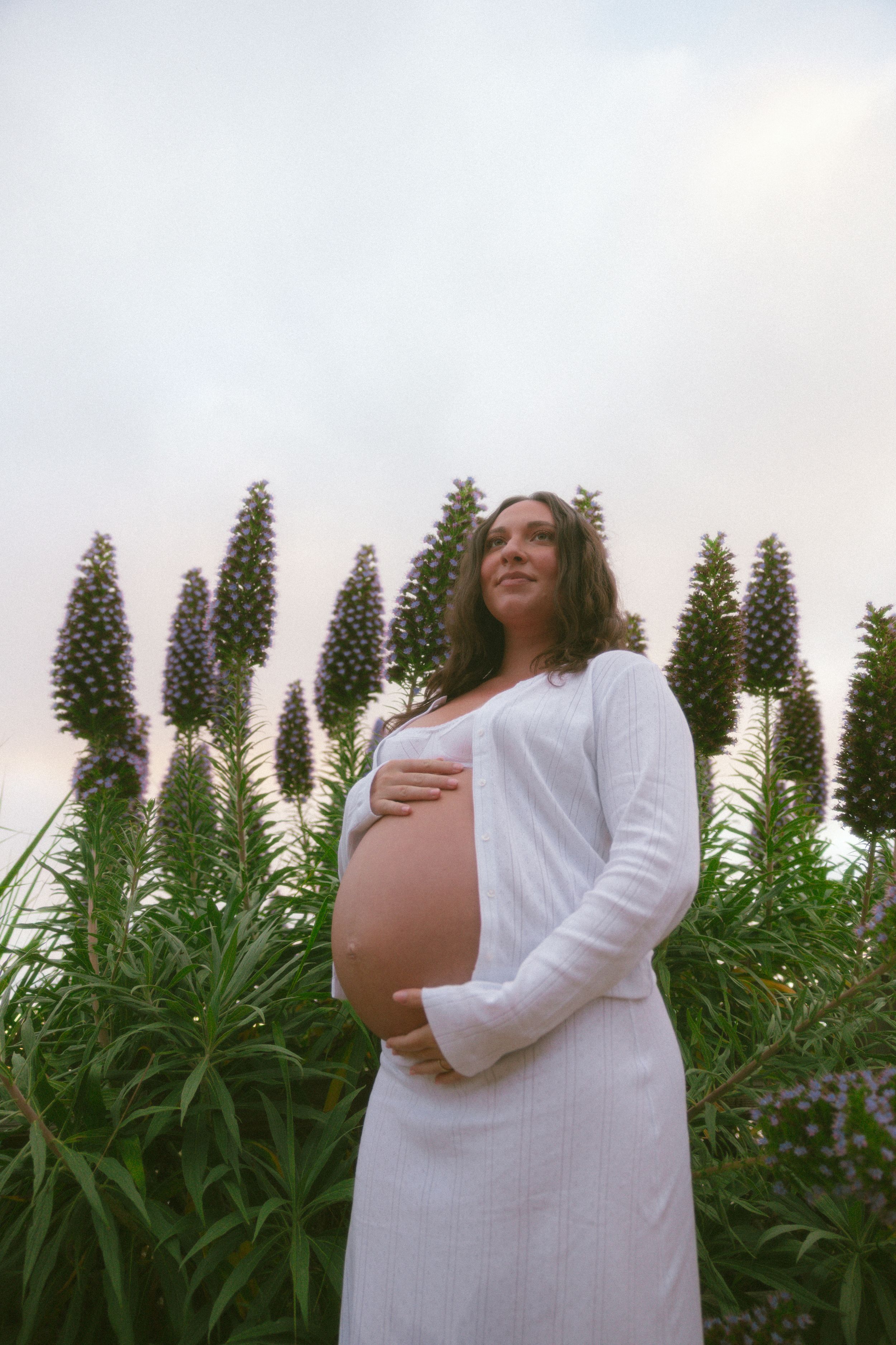 Pregnant woman in white dress holding baby bump, standing before tall purple flowering plants outdoors.