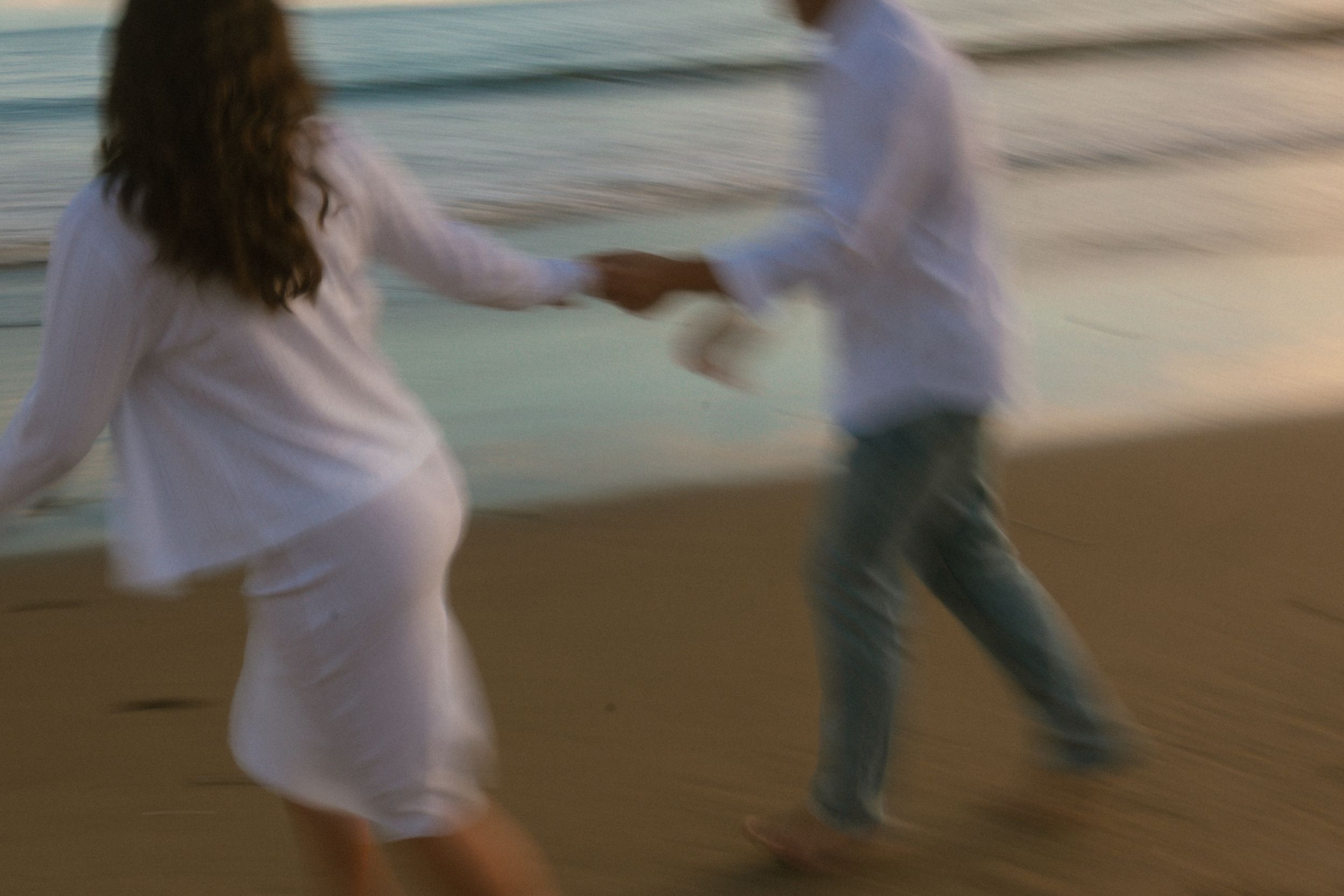 Couple holding hands dancing on a sandy beach at sunset, wearing white outfits in a romantic blurred motion photo.