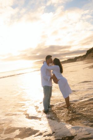 Couple sharing a romantic kiss on a sunlit beach at golden hour, waves washing over their feet.
