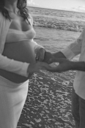 Couple holds hands on beach shoreline, pregnant woman in white dress with ocean waves in black and white.