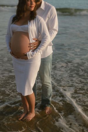 Pregnant couple standing barefoot on a beach, woman in white dress cradling baby bump, man embracing her from behind.