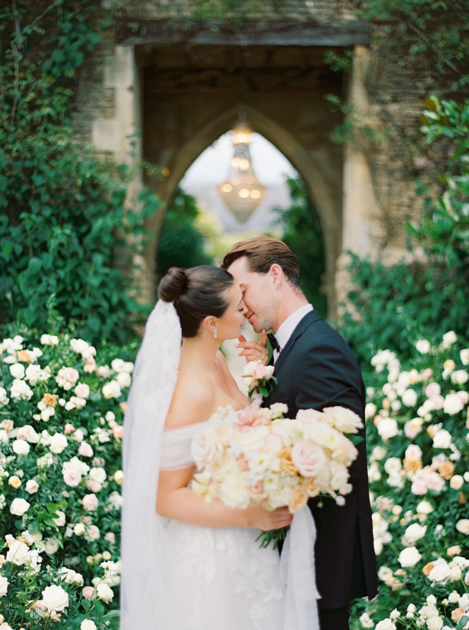 Bride and groom share a kiss surrounded by lush white roses in an elegant garden with a stone arch backdrop.
