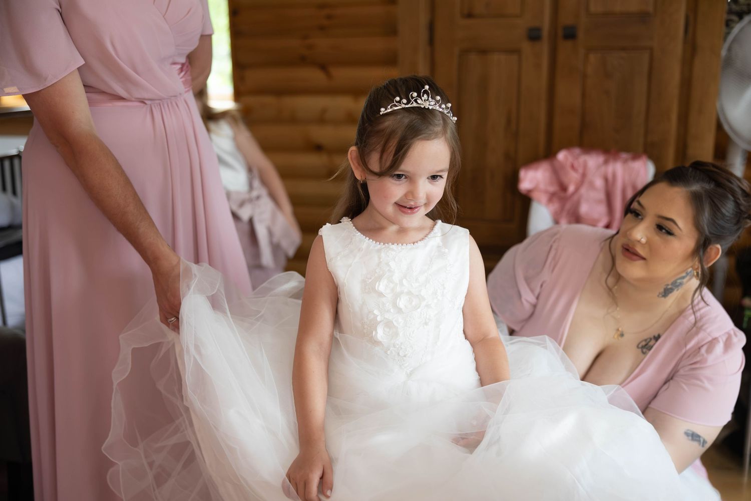 Adorable flower girl in white tulle dress with tiara gets dress adjusted by bridesmaids in pink gowns.