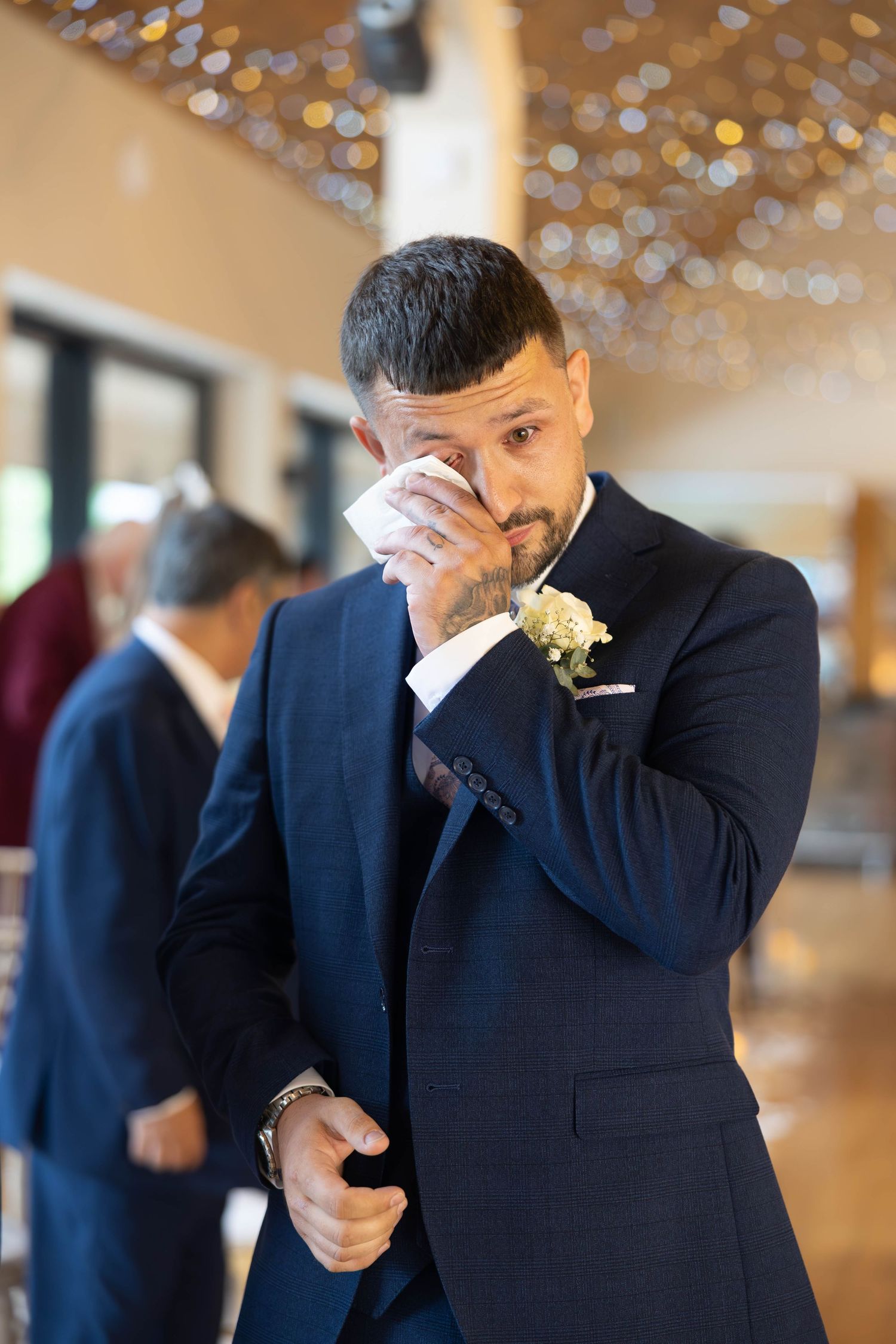 Emotional groom in navy suit with boutonniere dabs tears with handkerchief during wedding ceremony.